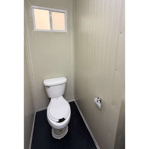 Interior de remolque sanitario 12x7 pies con sanitario instalado, paredes metálicas color beige y ventana superior para ventilación.