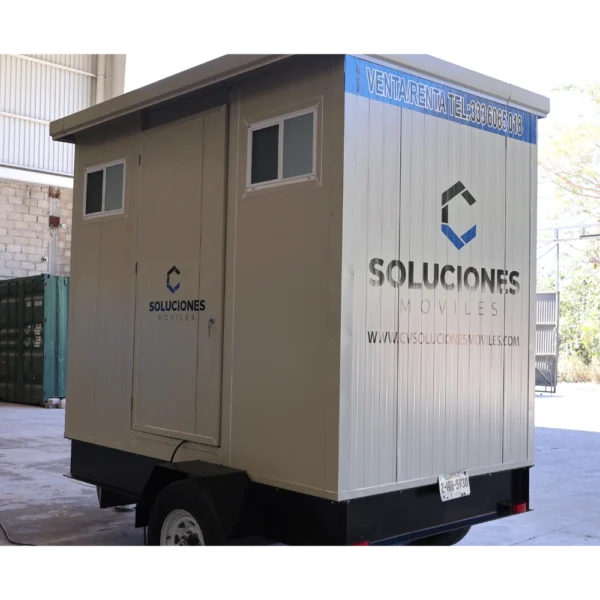 Remolque sanitario móvil de 12x7 pies color beige y gris con logotipo de Soluciones Móviles, diseñado para venta o renta, ubicado en un almacén industrial.